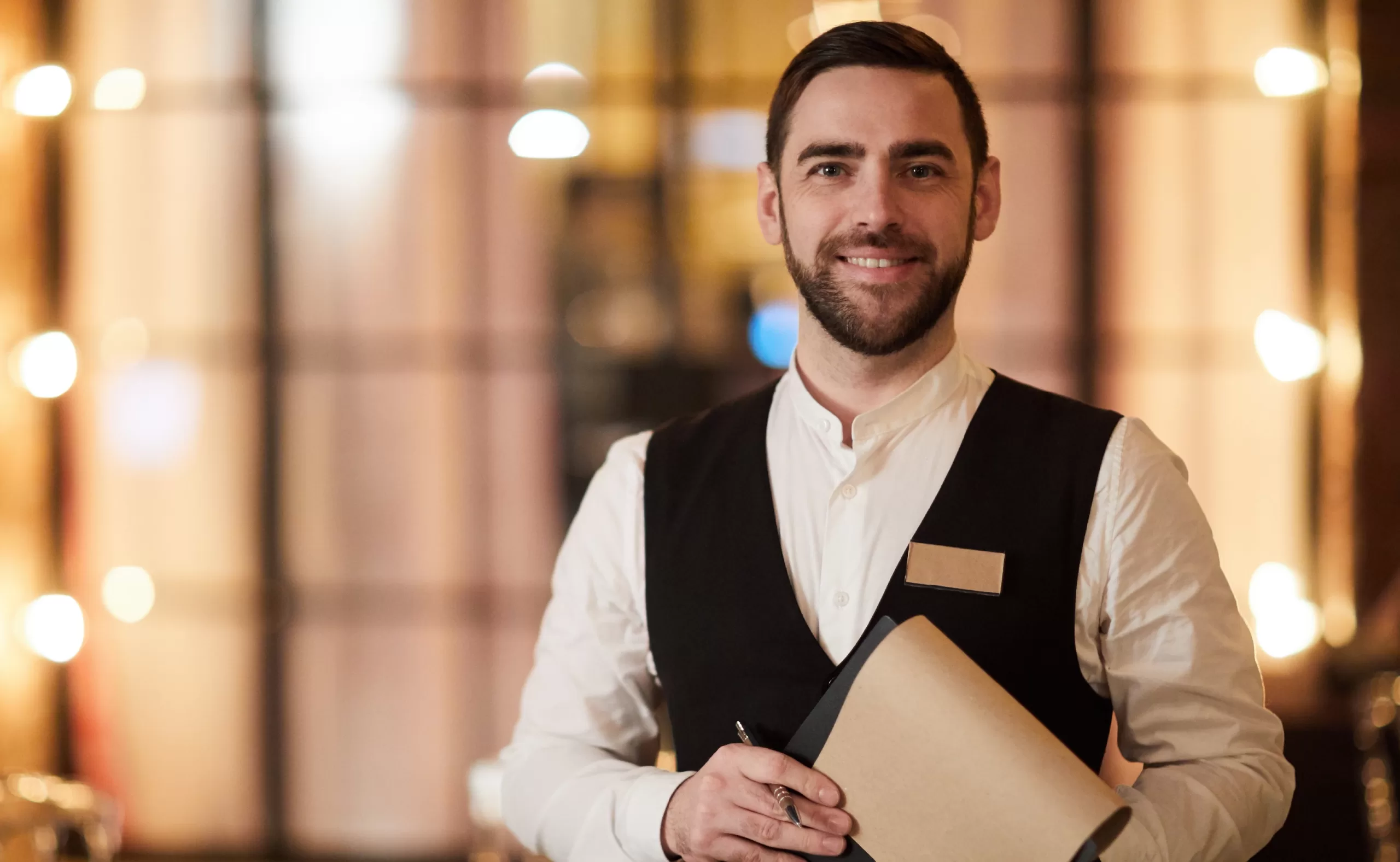 a skilled young man holds clipboard and wears hotel waiter uniform in a hotel restaurant