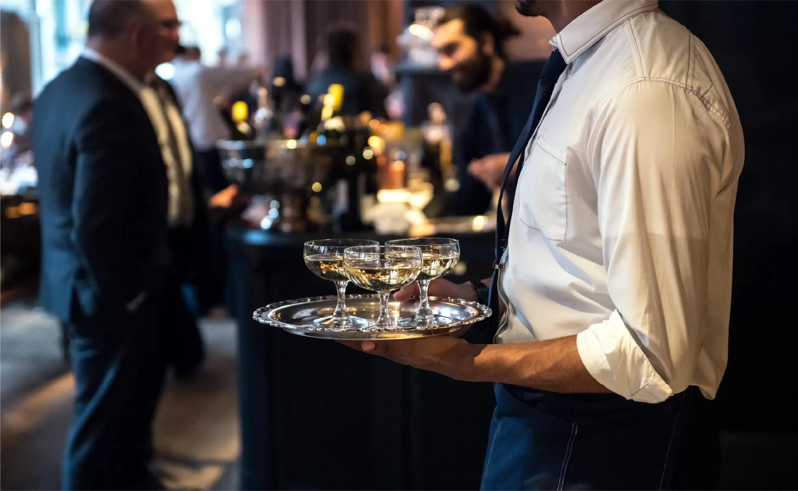 Waiter holding a tray of wine glasses at a formal event with people socializing in the background.