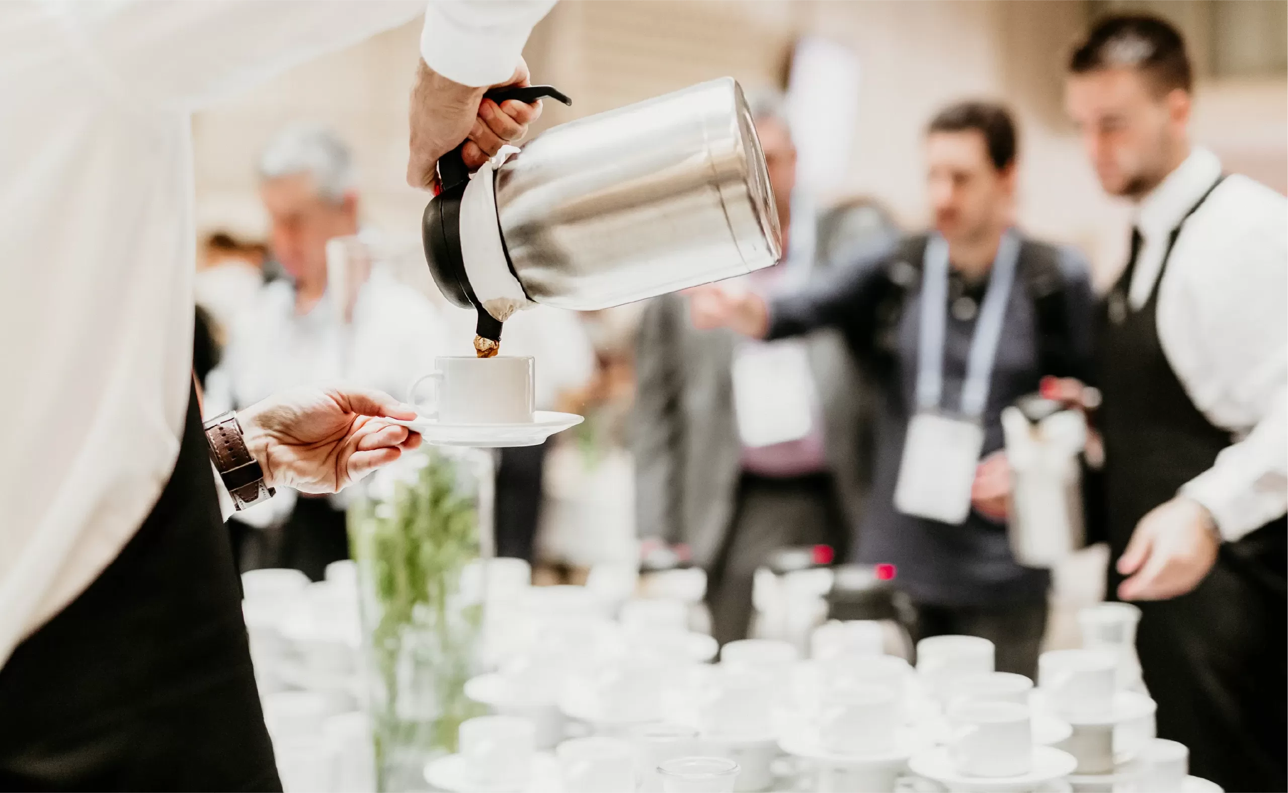 Server pouring coffee into a white cup at a busy catered event, with banquet staff and guests in the background.