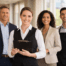 Four hospitality team members stand in a warm, sunlit hotel lobby corridor, with one holding a clipboard and a softly blurred city view outside.