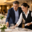 A supervisor coaches a server on table settings in an elegant banquet hall with warm ballroom lighting and softly blurred round tables in the background.