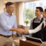 An area manager in business casual shakes hands with a front desk staff member in a bright hotel lobby, with a softly blurred Houston skyline visible through the windows.