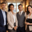 Four hospitality professionals stand in a boutique-hotel corridor with warm sconces and wood trim, one holding a clipboard in soft natural light.