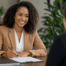 Two professional women in business attire collaborate at a modern reception desk, one holding a clipboard while the other speaks on a phone.