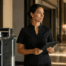 Hotel housekeeping manager holding a tablet beside a cleaning cart while reviewing staffing and turnover challenges in a modern hotel corridor.